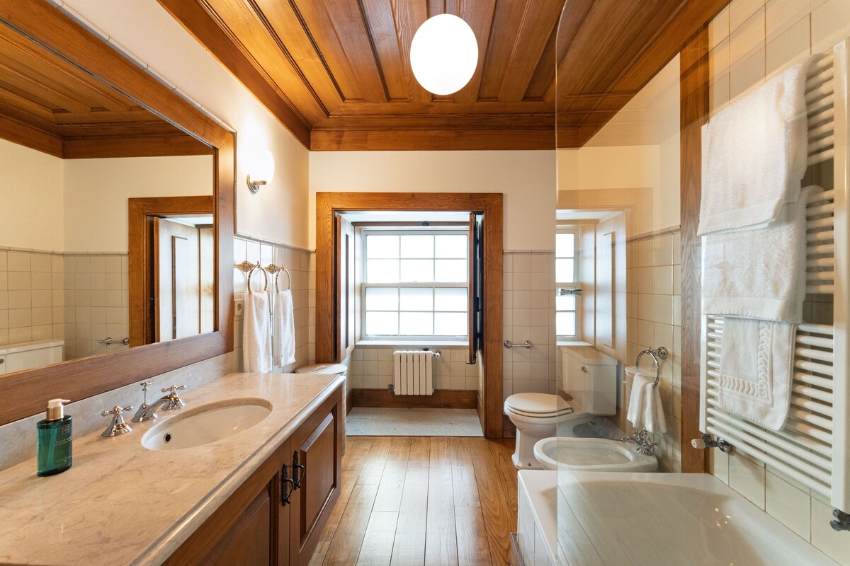 A bathroom with wooden ceiling beams, a large mirror, two sinks, a toilet, and a bathtub.