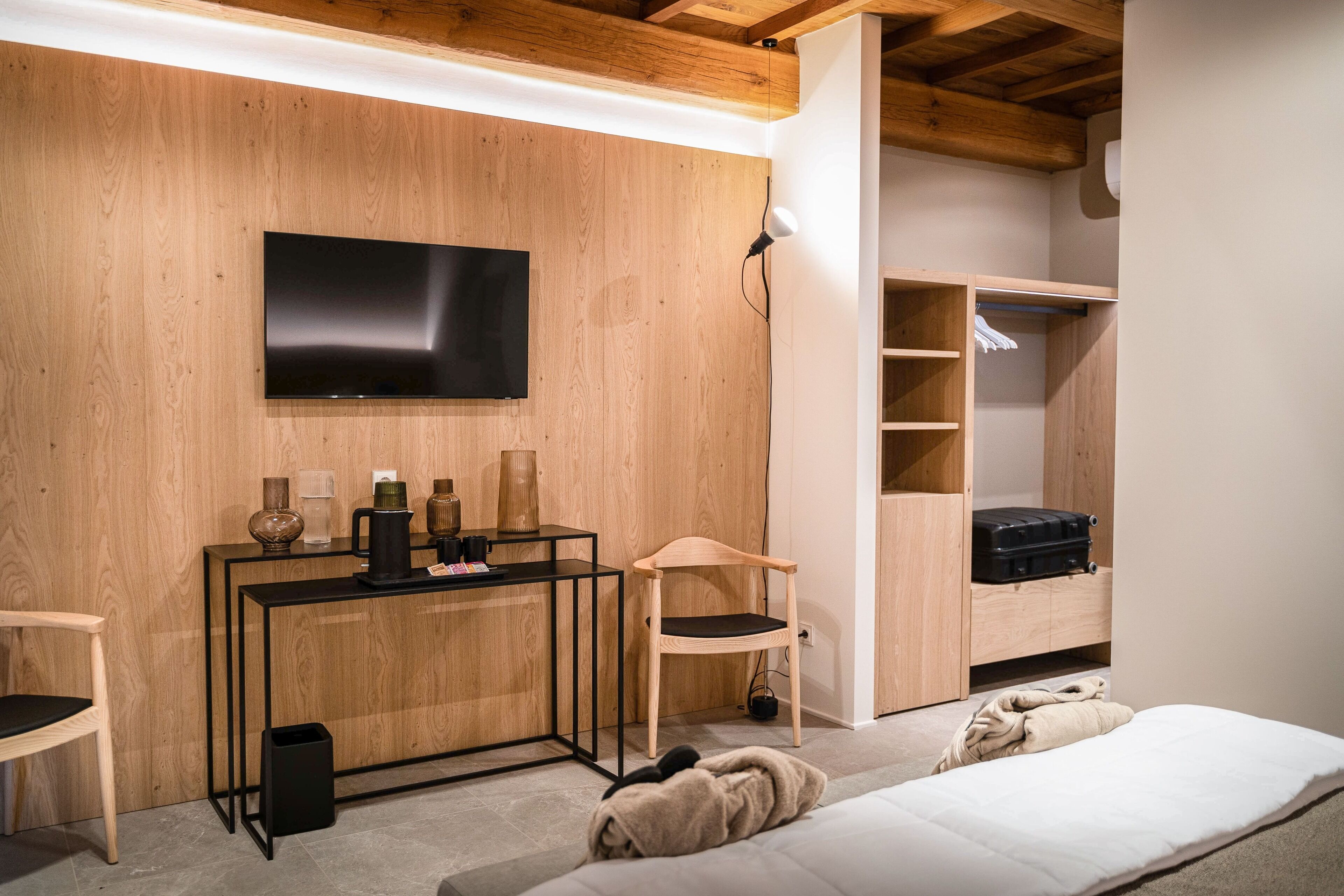 Una habitación moderna con una pared de madera, un televisor montado sobre una consola negra, una silla de madera, una cama y un armario empotrado con espacio para colgar.