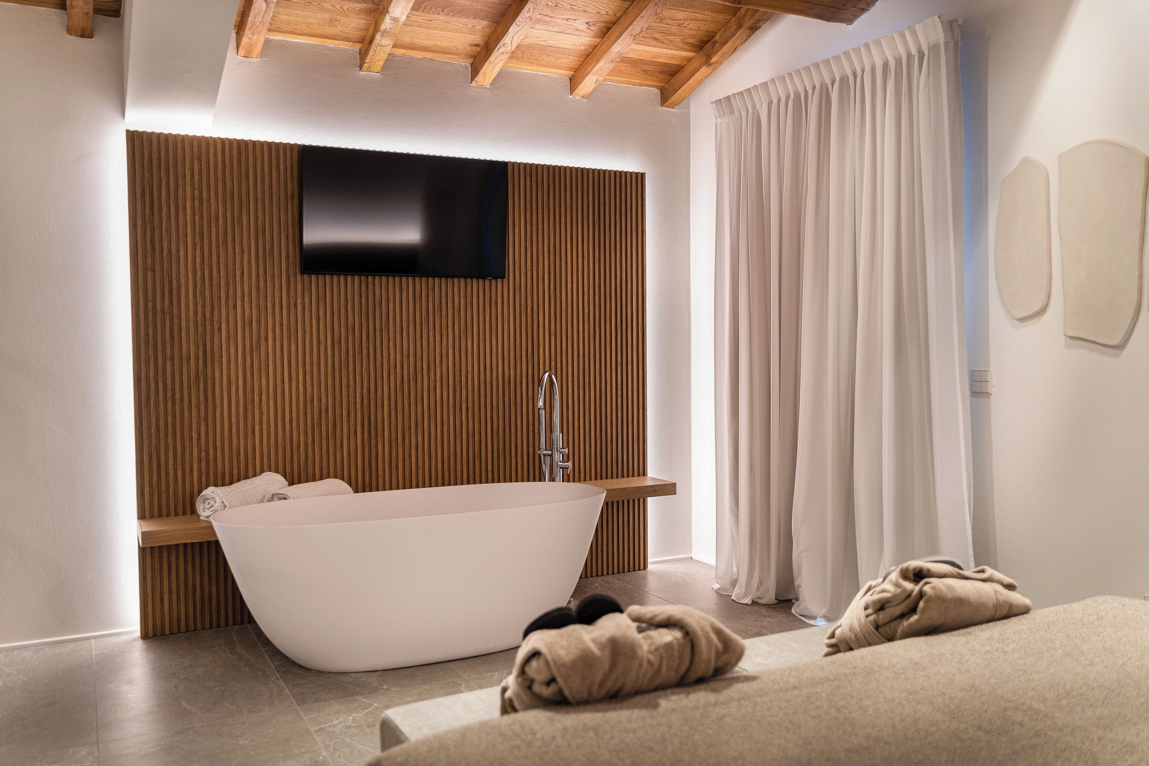A modern bathroom with a freestanding bathtub, wooden paneling, a mounted TV, and a large window with curtains.