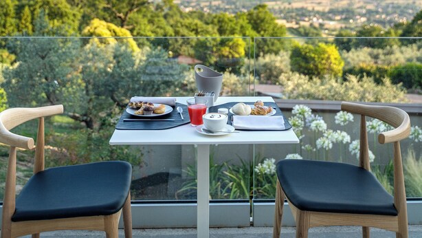 Una tavola imbandita con prodotti per la colazione e un bicchiere di succo di frutta rossa, con vista su un paesaggio pittoresco tra alberi e montagne lontane.