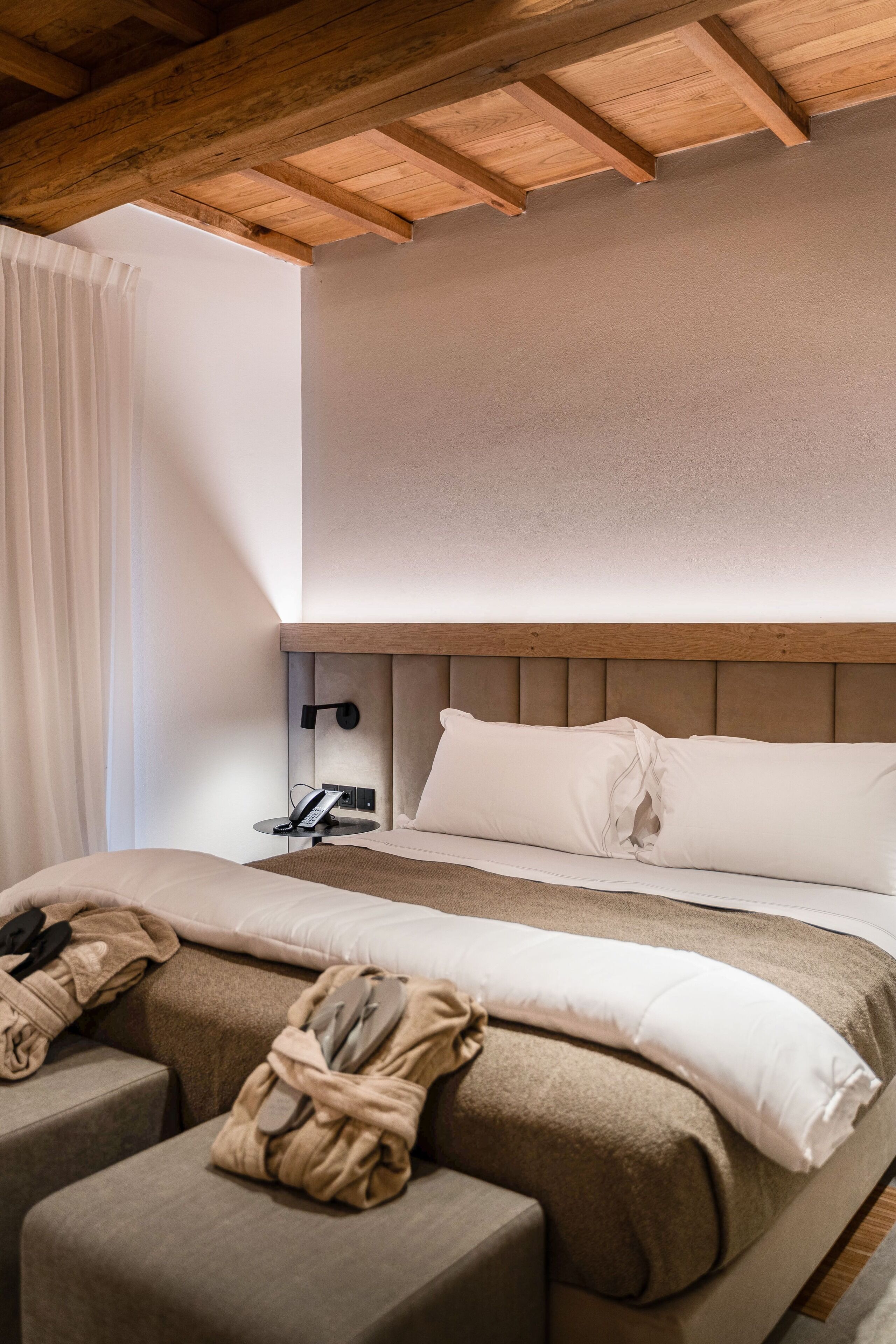 A modern hotel room with a wooden ceiling, a bed with white linens, a bedside table with a lamp, and a small seating area with a blanket.