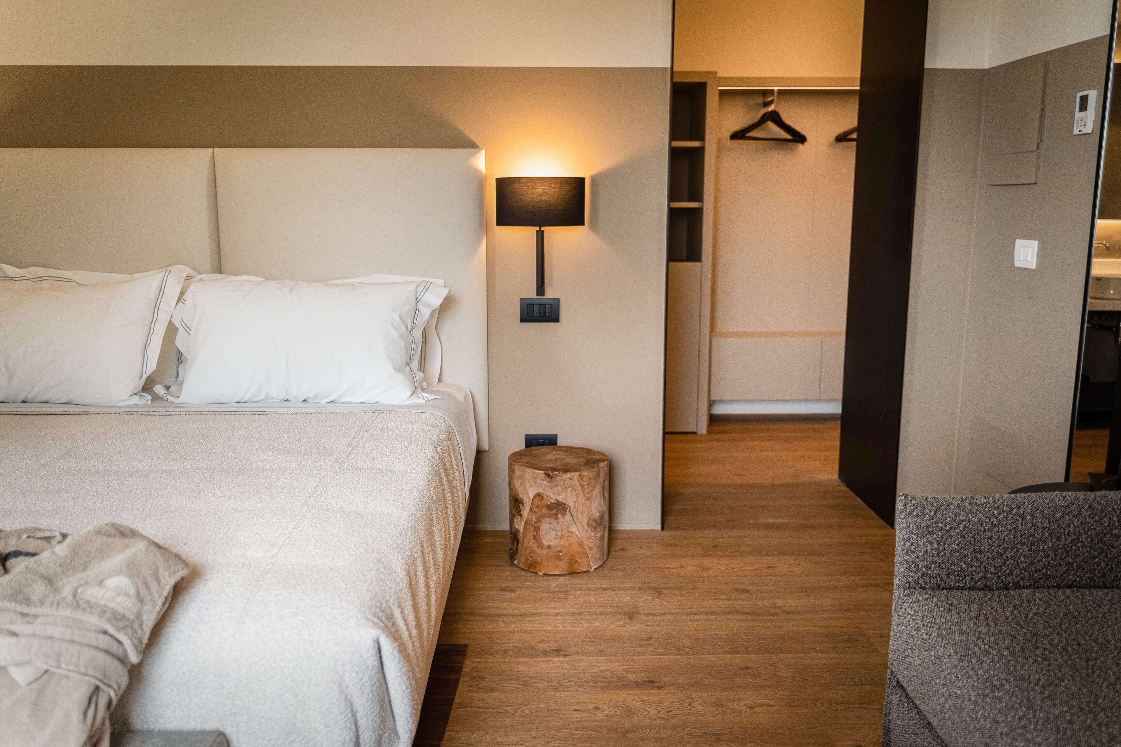 A modern hotel room with a large bed, a bedside lamp, a wooden stool, and a grey armchair.