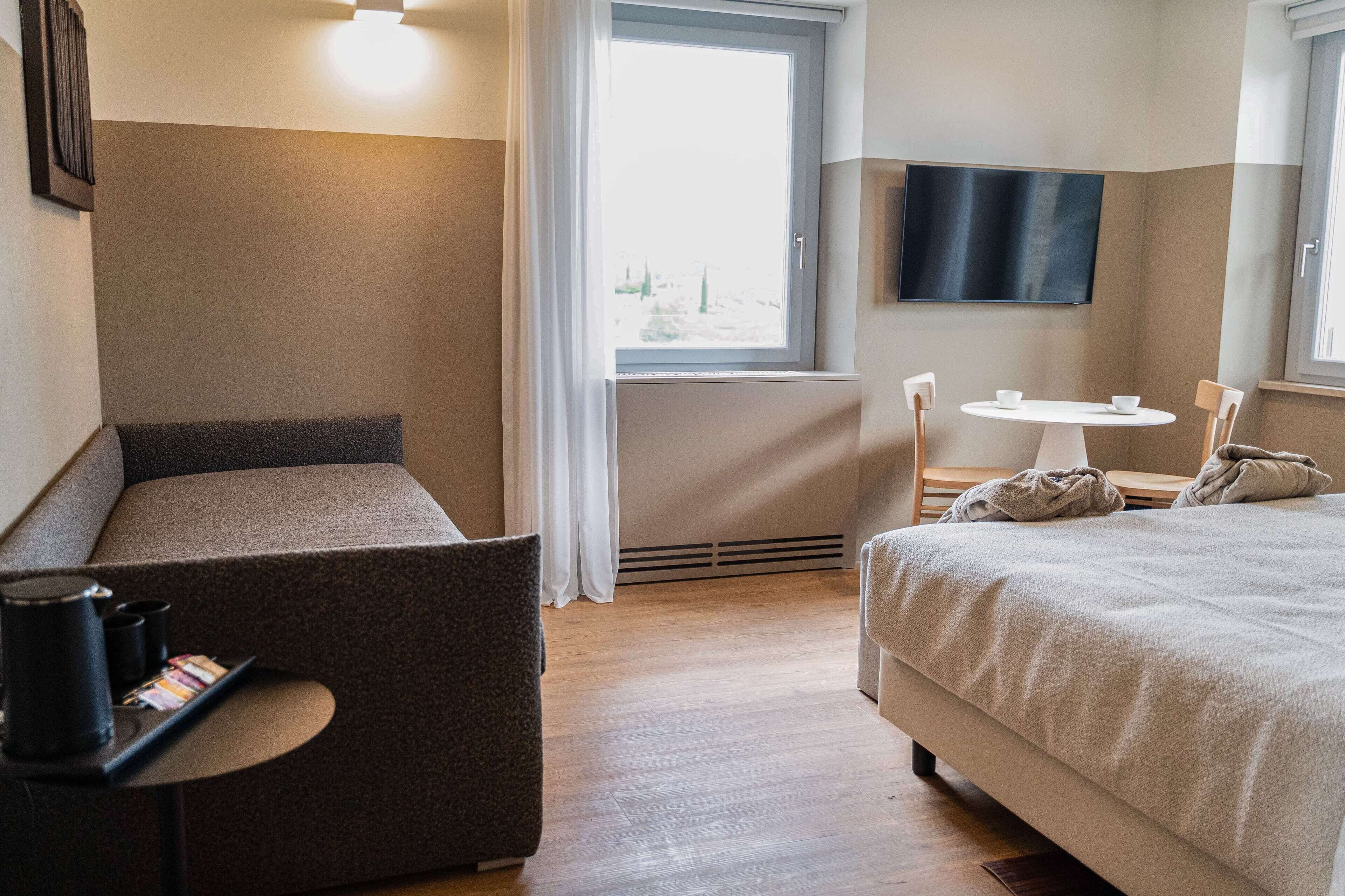 A hotel room with a bed, a seating area with a table, a flat-screen TV, and a window with a view of trees.