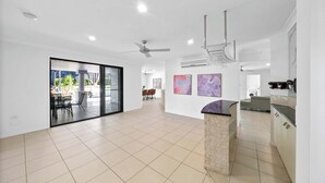 A spacious living area with a sliding glass door leading to an outdoor patio, a kitchen with a stone island, and a ceiling fan. - Haven by Khove (Pacific Pines)