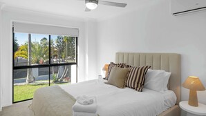 A modern bedroom with a large bed, a ceiling fan, a window with a view of palm trees, and a bedside lamp. - Haven by Khove (Pacific Pines)