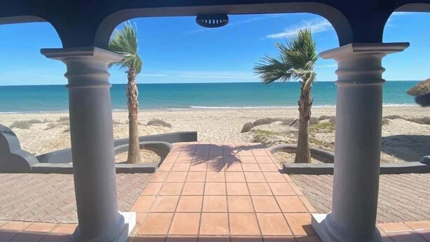 Un pasillo cubierto que conduce a una playa con palmeras y un cielo azul despejado.
