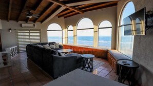 Amplia sala de estar con un sofá grande, una mesa de centro y un televisor montado en la pared. Varias ventanas arqueadas ofrecen vistas al océano.