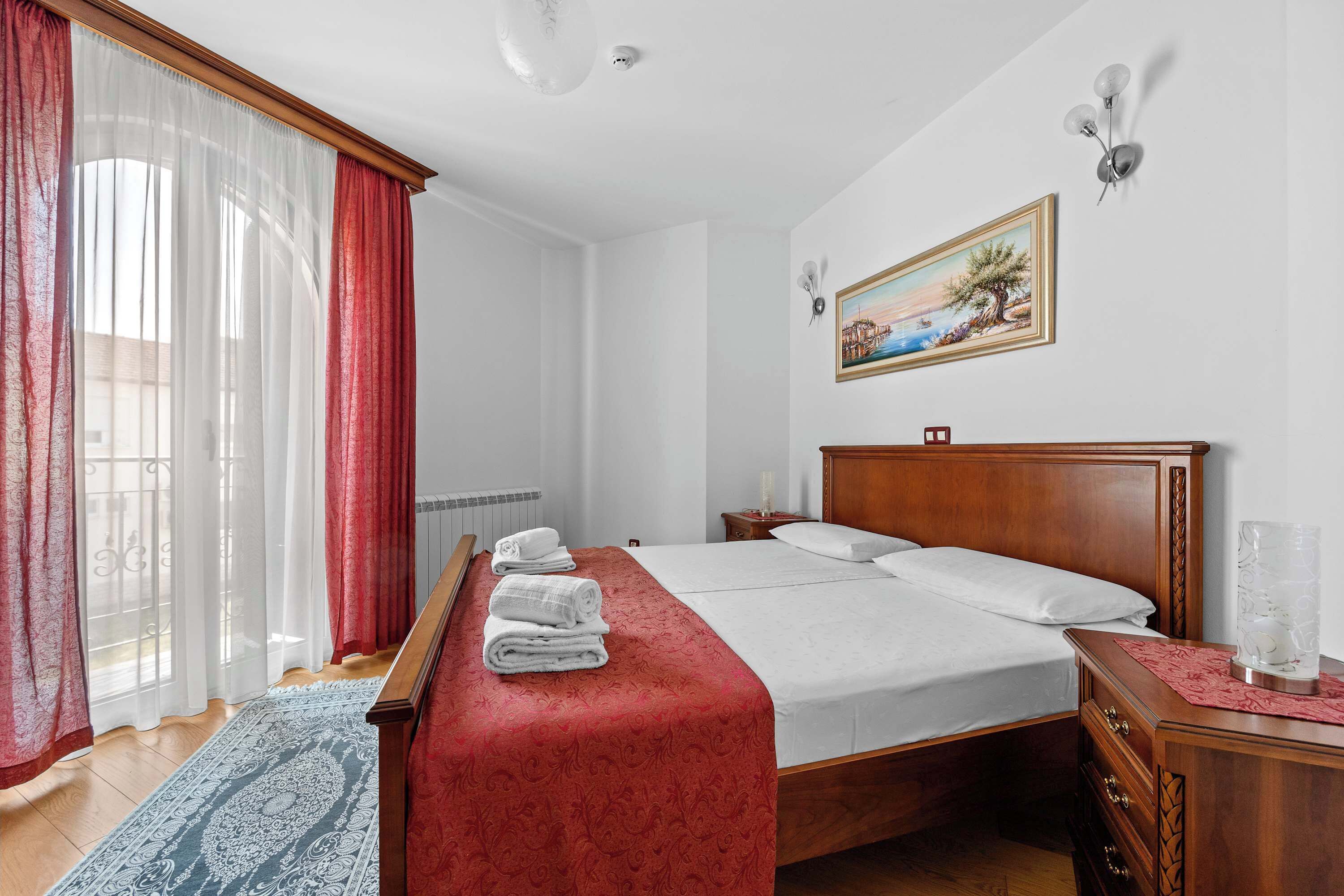 A hotel room with a large bed, a wooden nightstand, a painting on the wall, and a window with curtains.