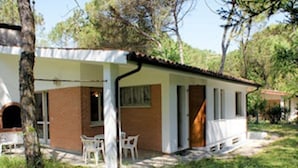 Uma casa térrea com fachada de tijolo, um alpendre com móveis brancos e uma pequena área de jardim com flores cor-de-rosa.