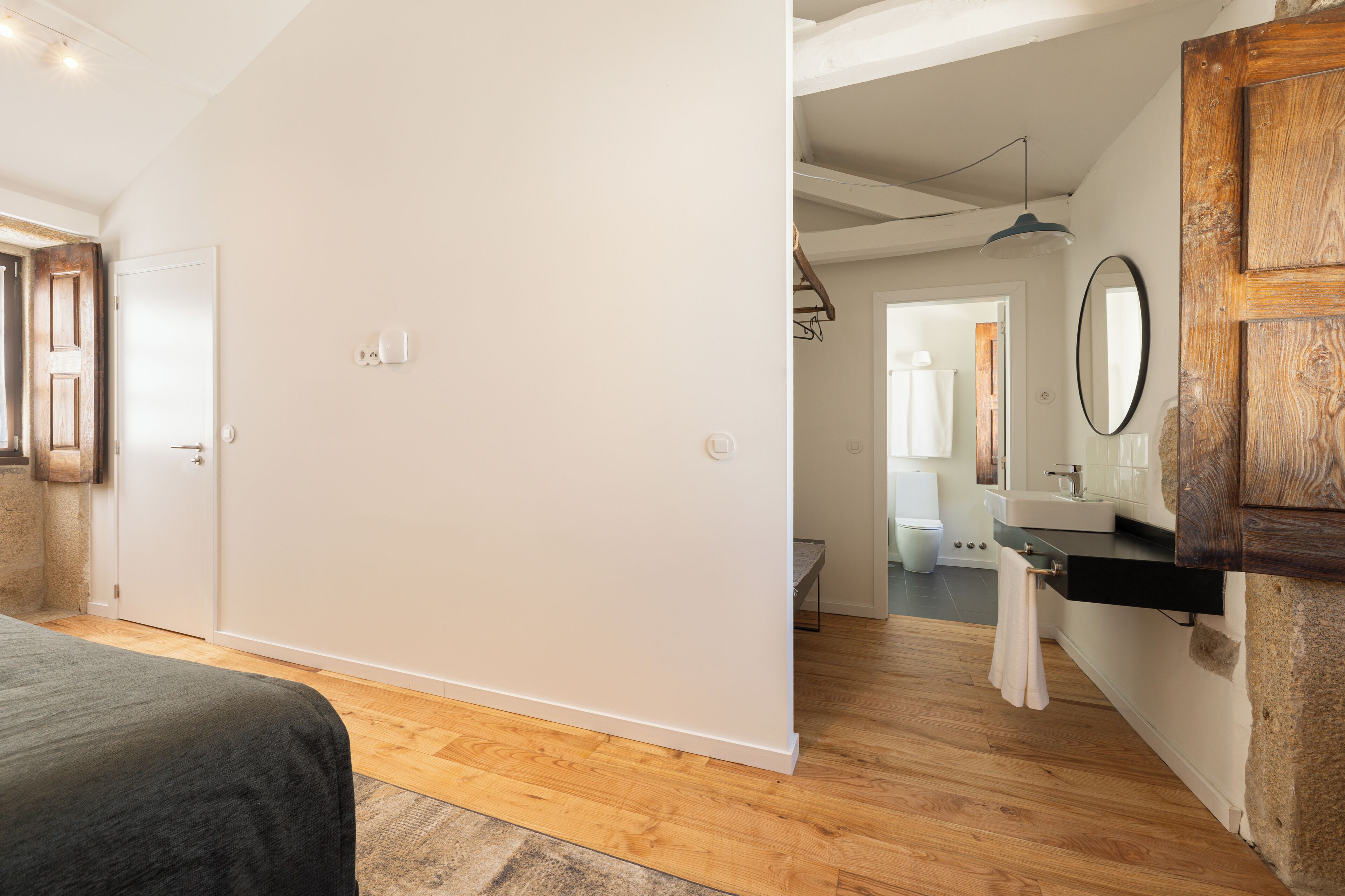 A bedroom with a bed, wooden flooring, a white wall, a wooden door, and a bathroom visible through an open door.