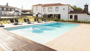 Una piscina con sillones y sombrillas, rodeada por una terraza de madera y un edificio blanco con tejado de baldosas.