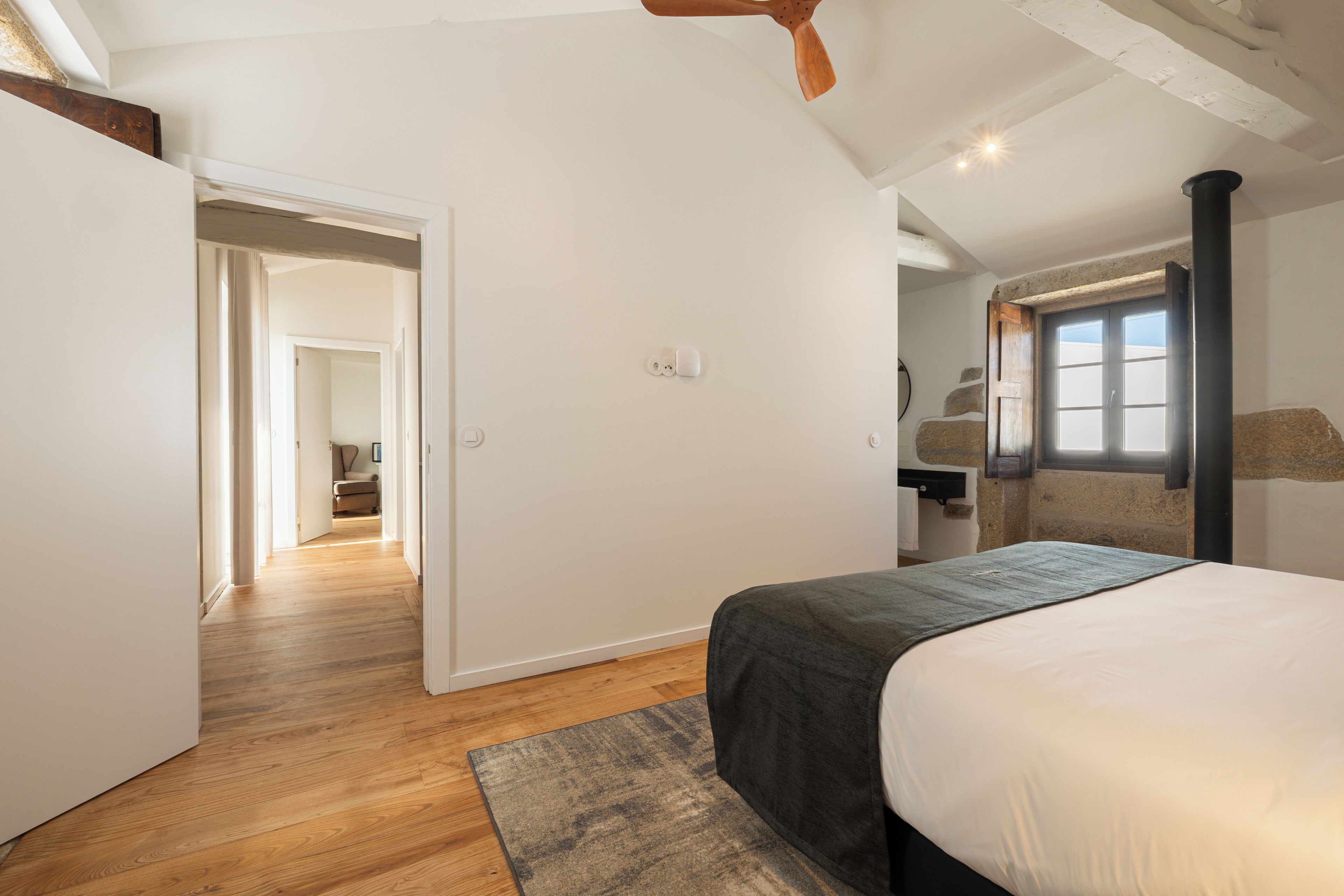 A bedroom with a bed, a wooden floor, a window, and a door leading to another room.