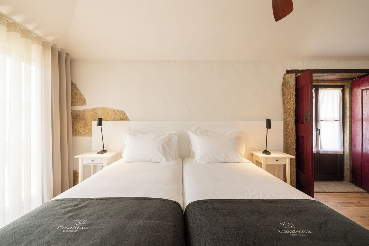 A double bed with white linens, two bedside tables with lamps, a wooden door, and a view of a room with a window and curtains.