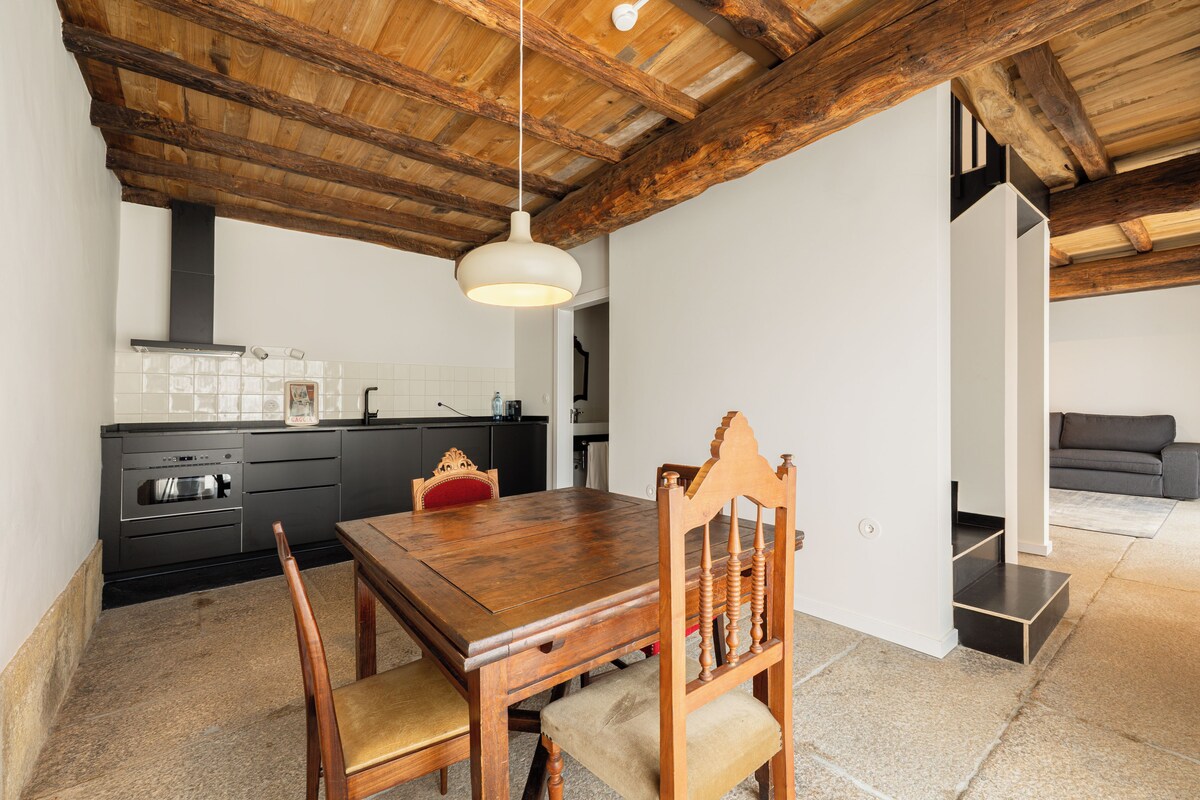 A dining area with a wooden table and chairs, a hanging light fixture, and a kitchen in the background.
