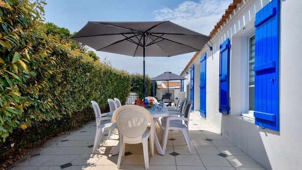 En udendørs terrasse med et bord og stole, en stor parasol og en hvid bygning med blå skodder.
