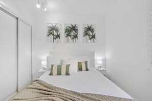 A neatly arranged bedroom with a large bed, white bedding, and three framed palm tree pictures on the wall.