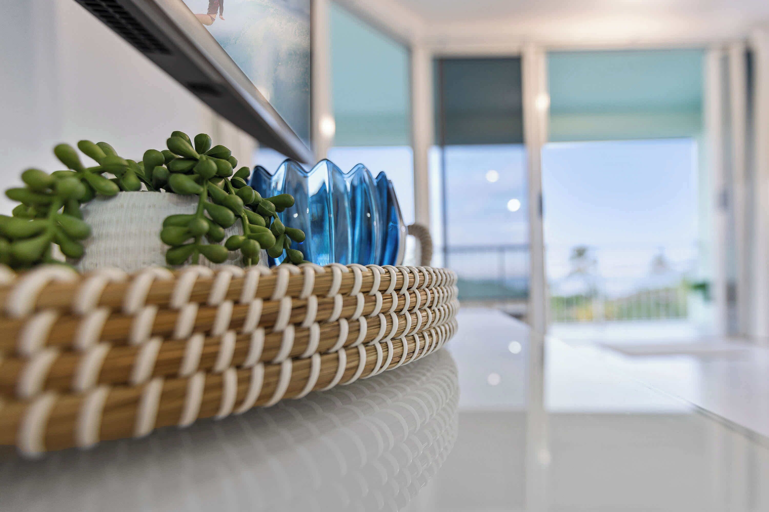 Un panier en osier contenant des plantes vertes et un vase bleu, le tout posé sur une surface blanche.