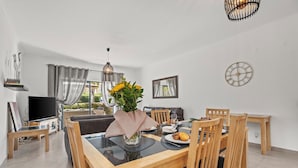 A modern dining area with a wooden table, chairs, and a vase of flowers. There is a television on a stand and a clock on the wall. - Coastal Charm Apartment (Lagos)