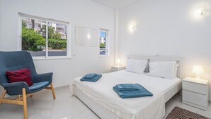 Ein Schlafzimmer mit Bett, Sessel, Nachttisch und Fenster mit Blick auf Gebäude und Grünflächen.