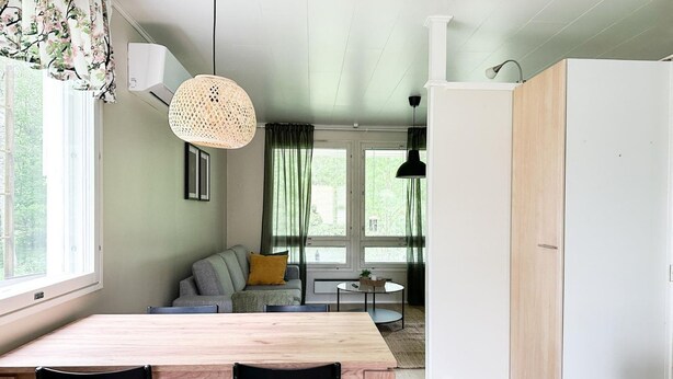 A modern kitchen with a wooden table and chairs, a hanging light fixture, and a view of a living area with a sofa and curtains.