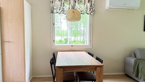 A dining area with a wooden table and chairs, a window with a view of greenery, and a wall-mounted air conditioner.