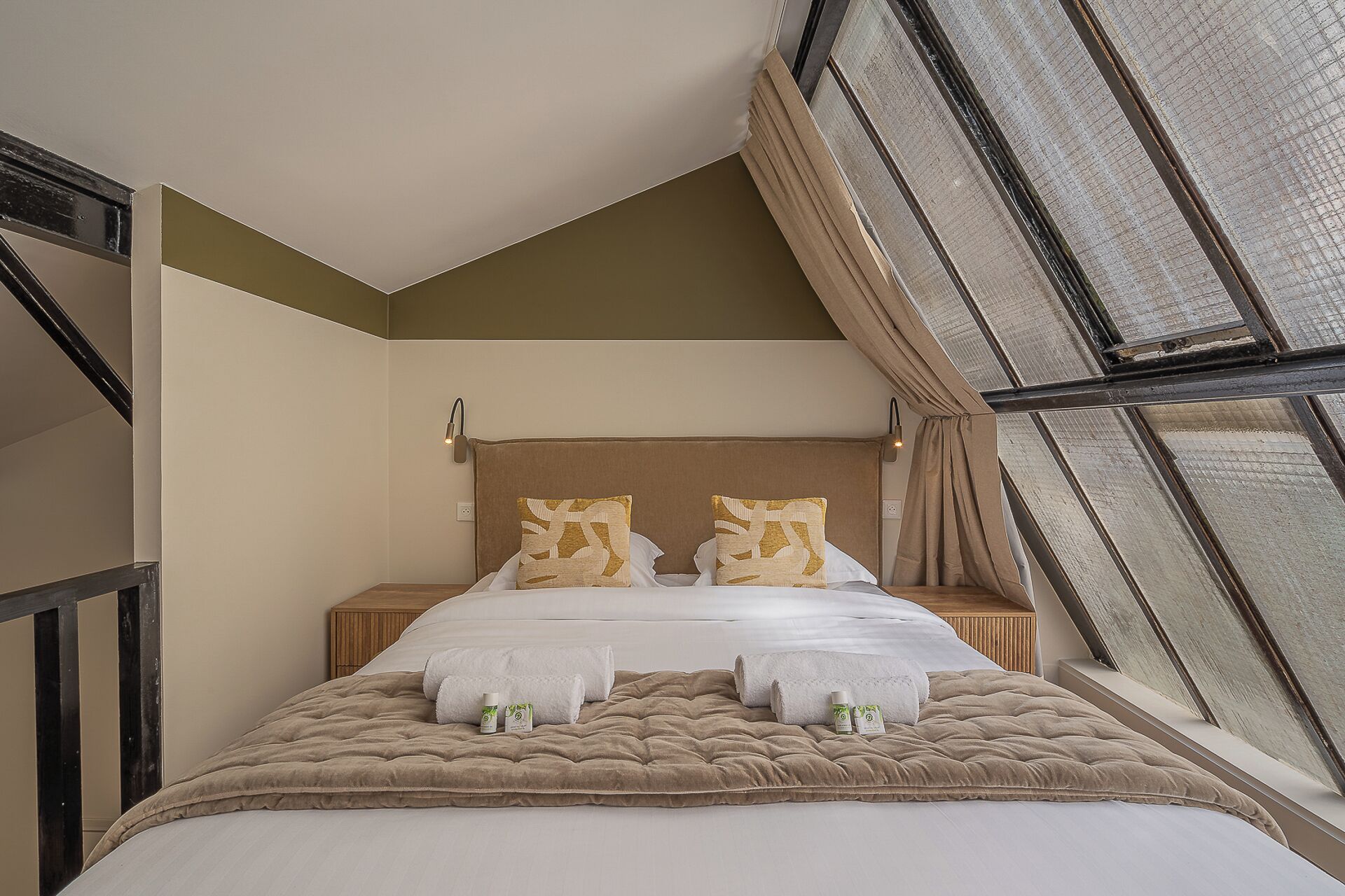 A bedroom with a large window, a bed with a beige quilt, two pillows, and a nightstand with a lamp.