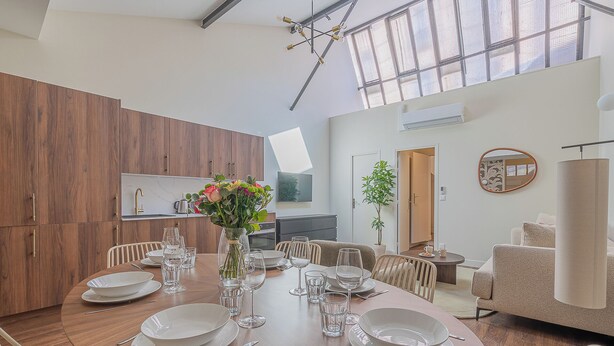 Een moderne keuken met houten kasten, een eethoek met een ronde tafel voor vier personen, en een groot raam dat natuurlijk licht binnenlaat.
