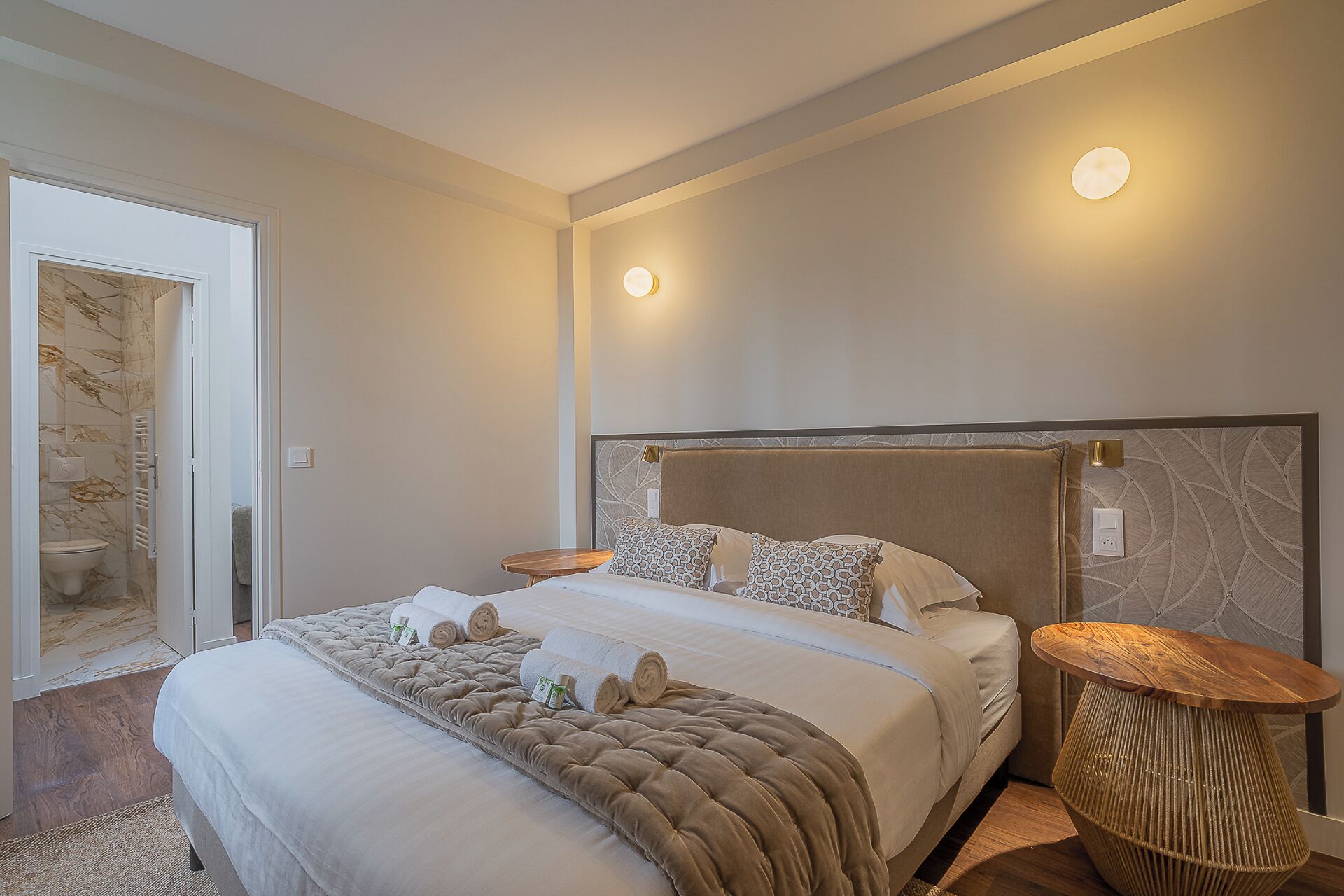 A modern hotel room with a large bed, two bedside tables, a headboard with a marble pattern, and a bathroom visible through an open door.