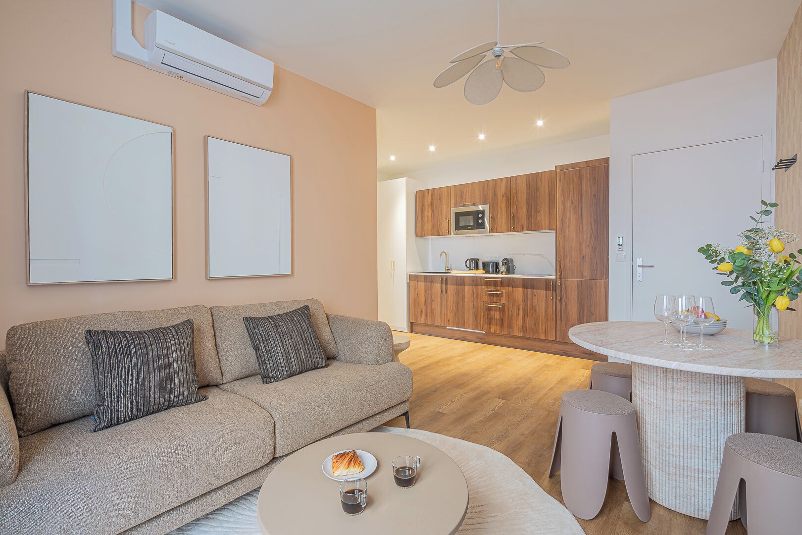 Un salon moderne avec un canapé, une table basse ronde sur laquelle se trouvent un croissant et des boissons, un coin repas avec une table ronde et des chaises, une cuisine avec des meubles en bois et un four à micro-ondes, et un ventilateur de plafond.