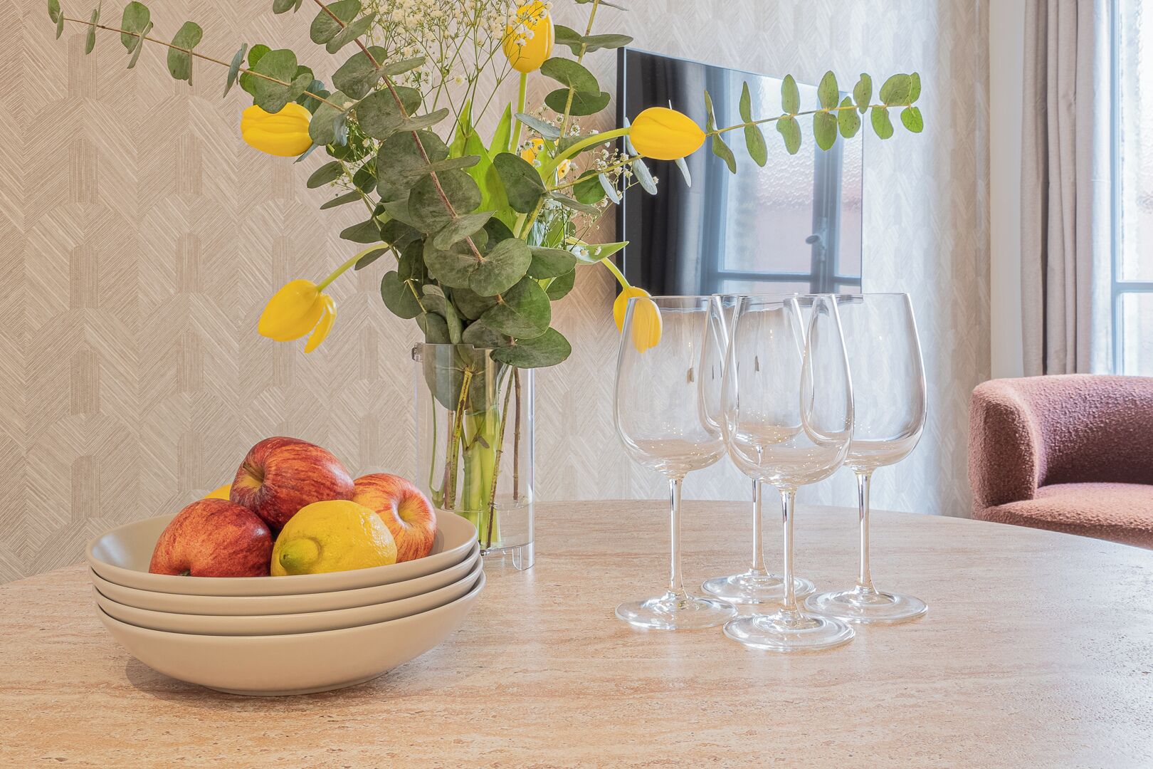 Uma mesa com uma tigela de frutas, um vaso de flores e copos de vinho vazios.