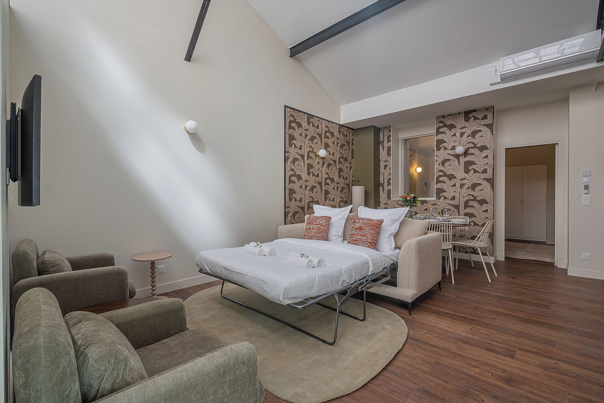 A modern hotel room with a bed, two armchairs, a small table, and a dining area with chairs.