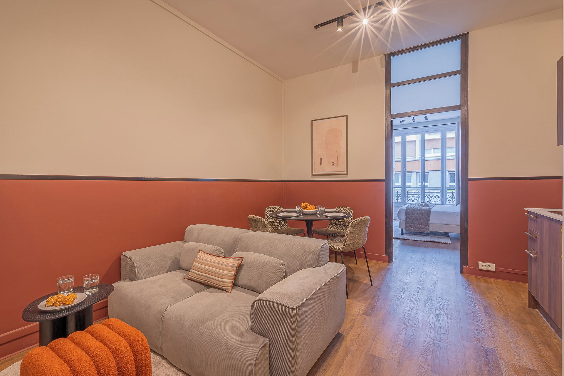 A living room with a grey sofa, a small table with glasses, and a dining area with patterned chairs.