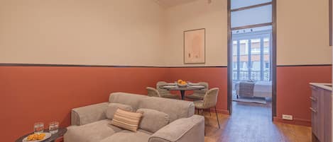 A living room with a grey sofa, a small table with glasses, and a dining area with patterned chairs.