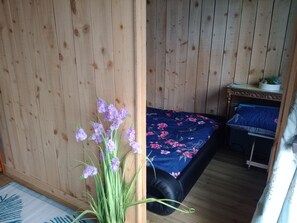 A room with wooden walls, a bed with a floral bedspread, a small table with a plant, and a window with a curtain.