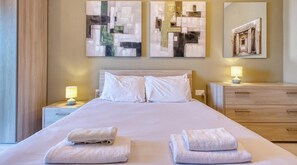 A hotel room with a bed, two bedside lamps, a wooden dresser, and abstract art on the walls.