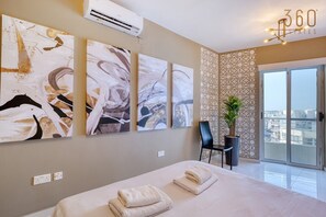 A hotel room with a bed, artwork on the wall, a chair, a potted plant, and a view of the city.