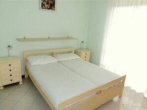 A bedroom with a wooden bed, two bedside tables, a nightstand, a wall-mounted shelf, and a single wall-mounted lamp. - Comfortable Apartment Near the Beach With Pool (Lignano Sabbiadoro)