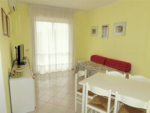 A dining area with a white table and chairs, a red sofa, and a window with sheer curtains. - Comfortable Apartment Near the Beach With Pool (Lignano Sabbiadoro)