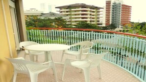 Un balcón con mesa y sillas, barandilla blanca y vista a edificios y follaje al fondo.
