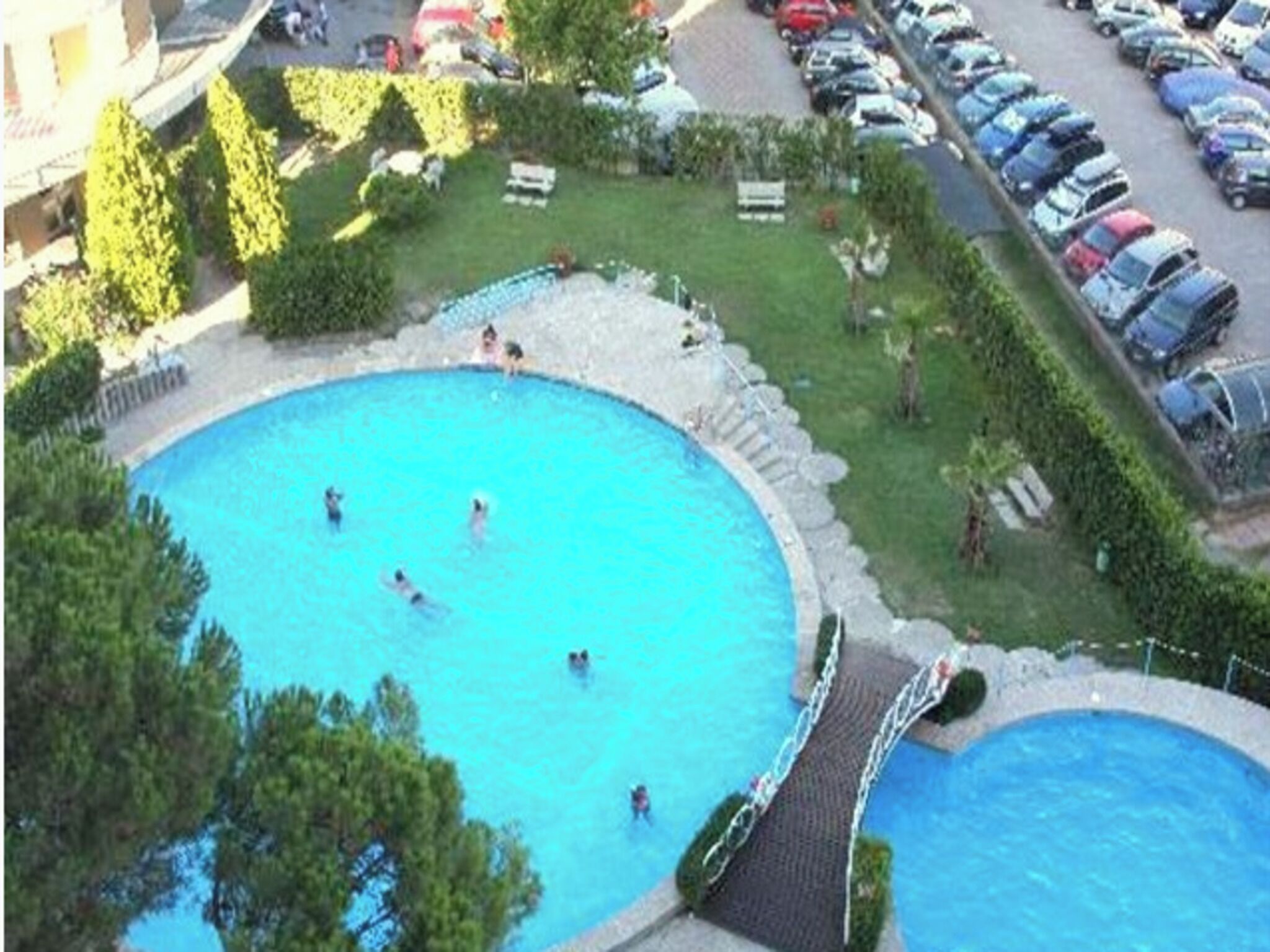A circular swimming pool surrounded by a stone deck, with a parking lot and greenery in the background.