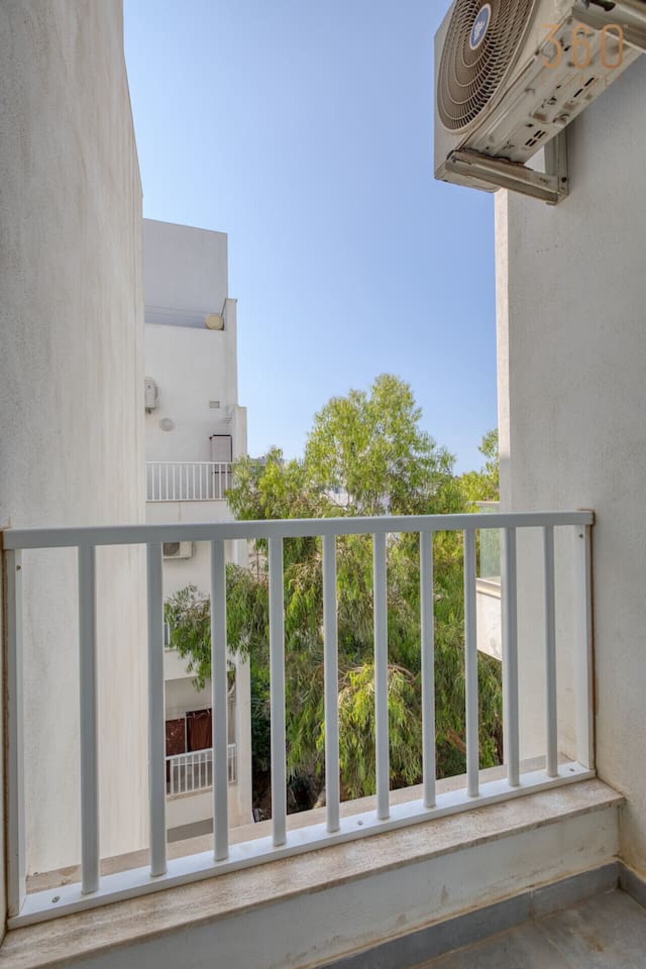 A balcony with a white railing, overlooking a residential area with trees and buildings.
