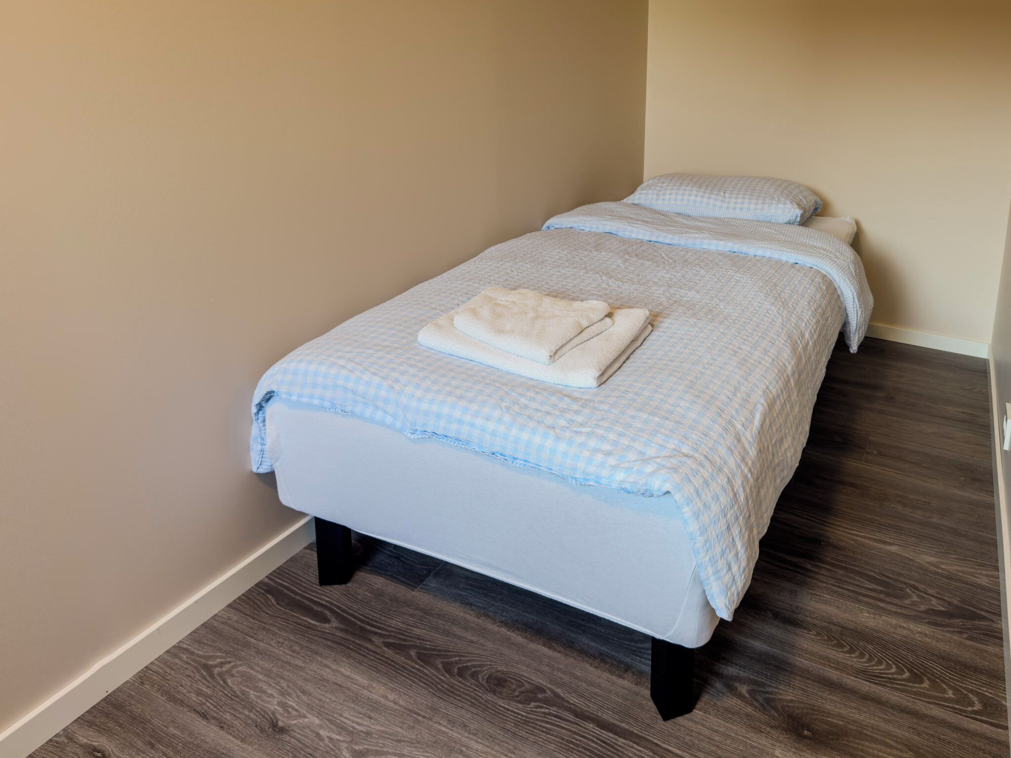 A small, neatly made bed with a checkered bedspread and two pillows, a folded towel on the bed, and a dark wooden floor.