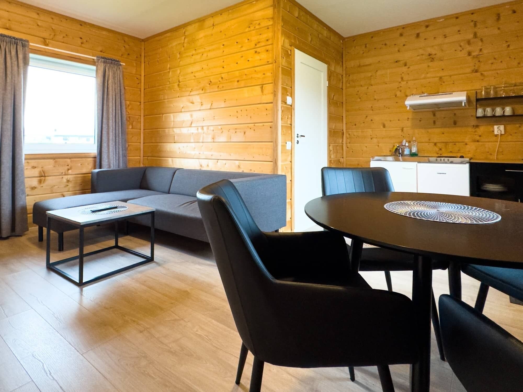 Une cabane en rondins avec un canapé, une table à manger avec des chaises, un coin cuisine et une fenêtre avec des rideaux.