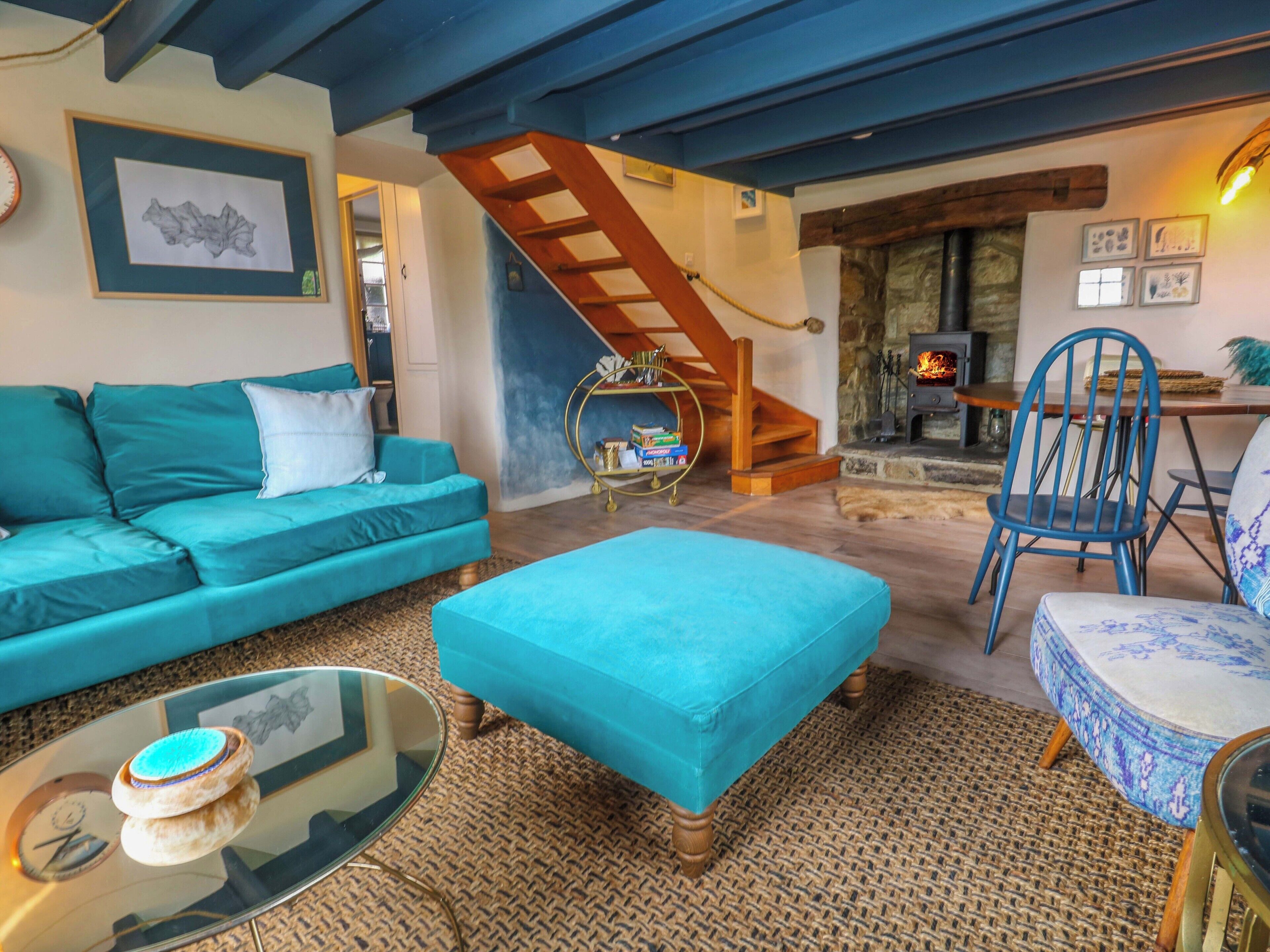 A cozy living room with a teal sofa, a matching ottoman, a glass coffee table, and a blue chair. A wooden staircase leads to a stone fireplace.
