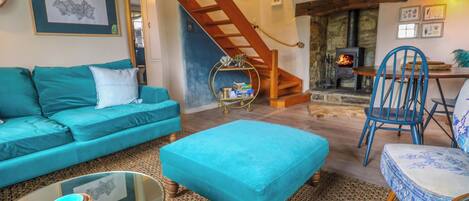 A cozy living room with a teal sofa, a matching ottoman, a glass coffee table, and a blue chair. A wooden staircase leads to a stone fireplace.