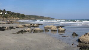 Una playa con grandes rocas, cielo despejado y casas al fondo.