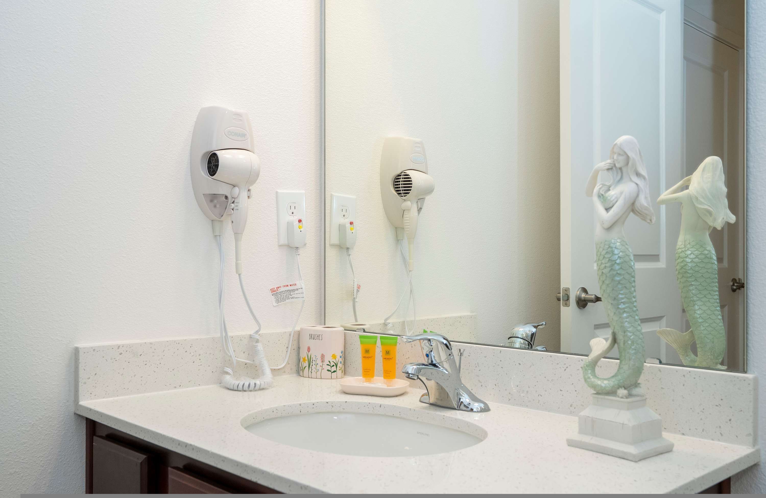 A bathroom with a sink, two hair dryers, a mirror, and a decorative mermaid statue.