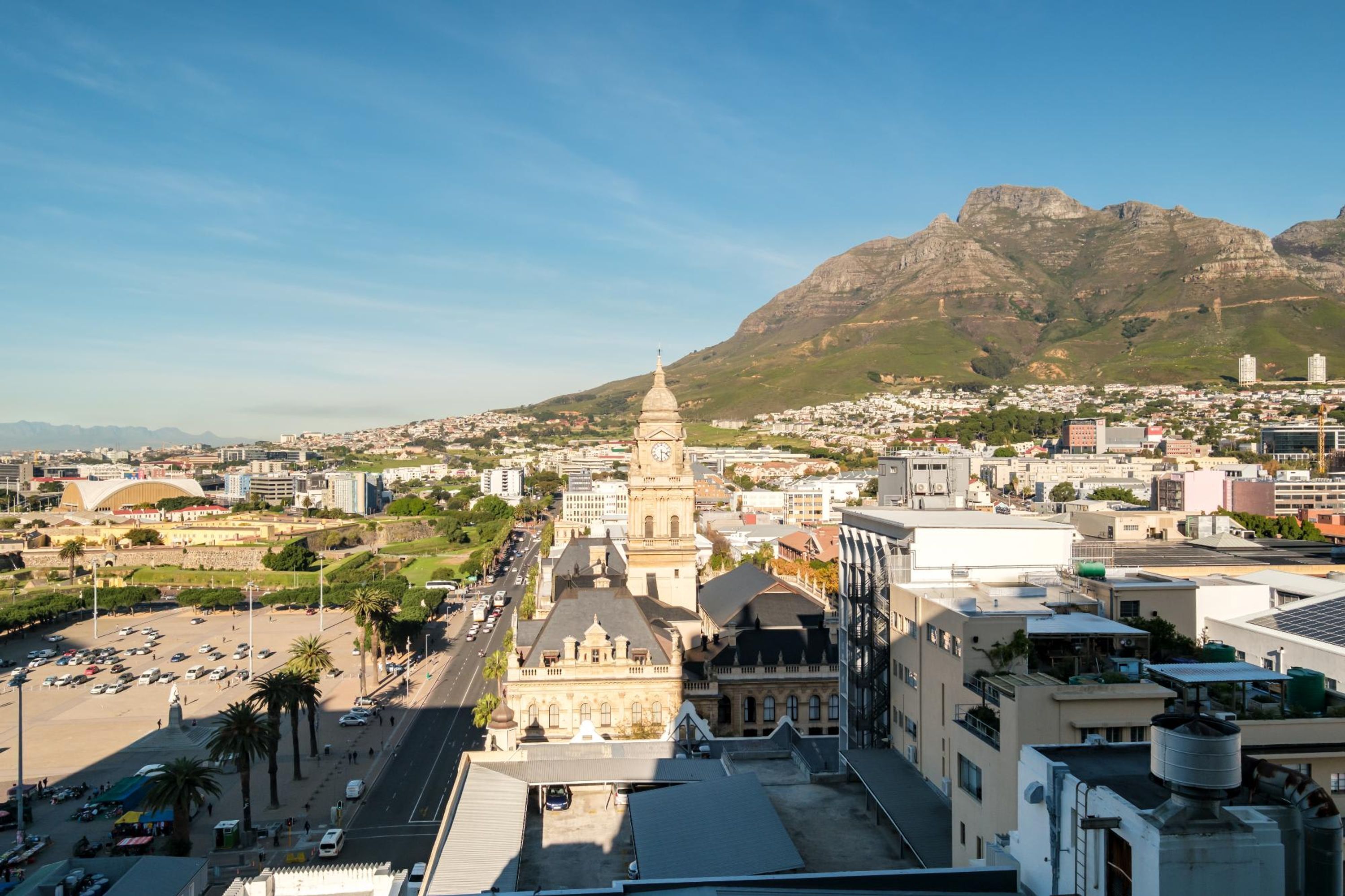 A cityscape with a prominent historical building featuring a clock tower, surrounded by modern buildings and a mountainous backdrop.