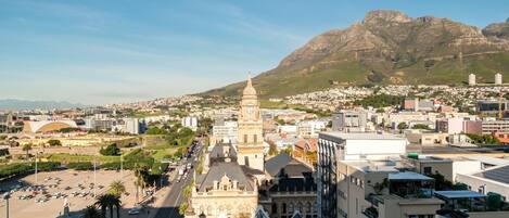 A cityscape with a prominent historical building featuring a clock tower, surrounded by modern buildings and a mountainous backdrop.