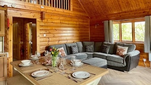 A wooden cabin interior with a dining table set for four, a grey sectional sofa, and a large window with a view of greenery. - Riverside Lodge—3-Bed Retreat in Aberdeenshire
Cozy, scenic, and peaceful. (Huntly)